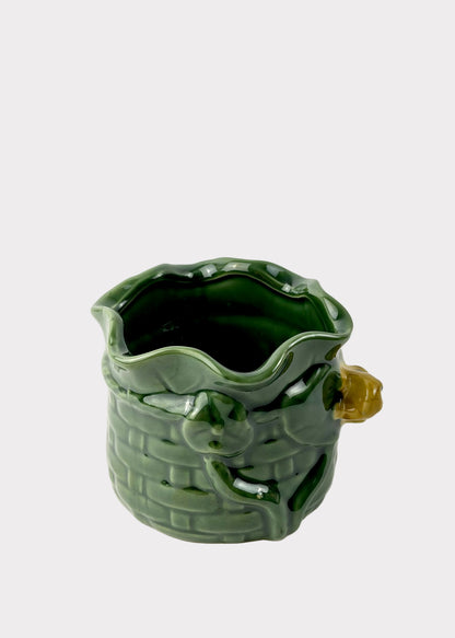 A green glazed ceramic pitcher shaped like a basket with a small yellow-green handle, photographed on a white surface against a white background.
