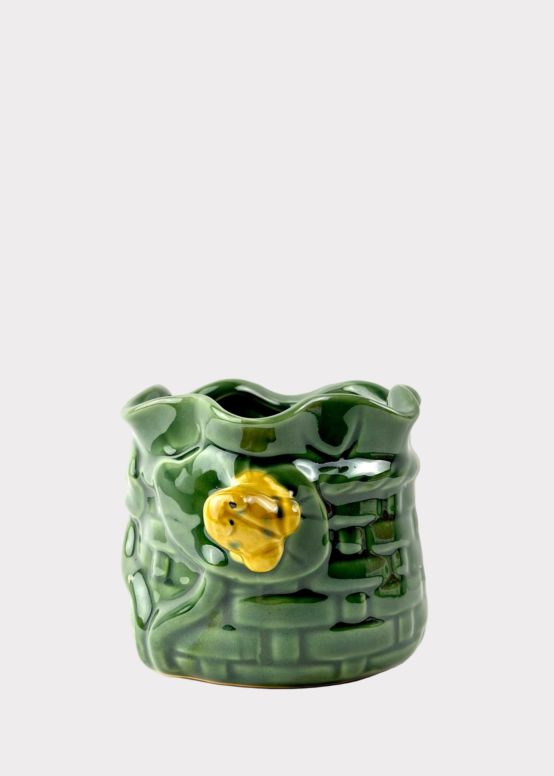 A green glazed ceramic planter shaped like a textured brick wall with a small yellow flower accent on the front, set against a plain white background.