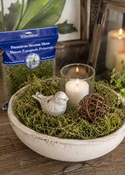 Decorative bowl with preserved Spanish moss, candle, bird figurine, and decorative ball on a wooden surface.