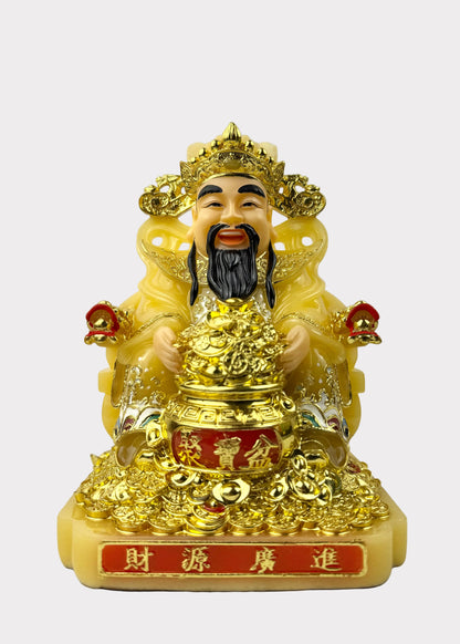 A golden Chinese deity figurine seated on a throne, holding a large gold ingot, with red accents and Chinese characters on the base, photographed against a neutral light gray background.