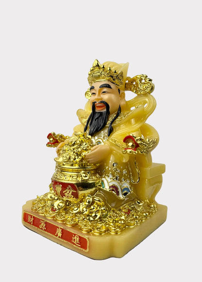 A golden Chinese deity figurine, likely representing Wealth (Fu Kun), seated on a base with Chinese characters. The figurine is primarily gold with a pale yellow body, a black beard, and small red accents.