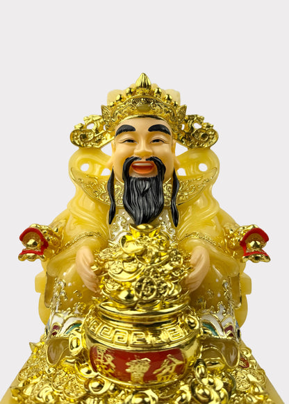 A golden Chinese deity figurine, likely representing Wealth or Good Luck, seated on a throne and holding a large gold ingot. The figure has a cream-colored face and black beard, wearing a yellow robe with red accents, set against a plain light gray background.