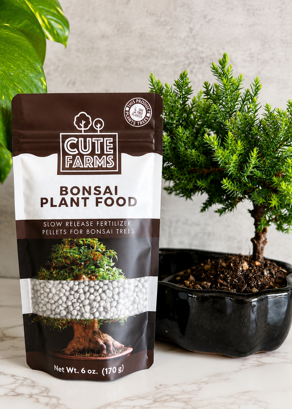 A 6 oz pouch of Cute Farms Bonsai Plant Food standing on a marble surface next to a small potted bonsai tree.