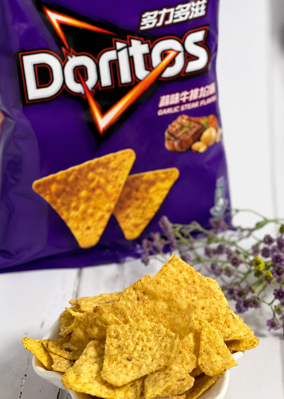 A bag of Doritos Garlic Steak Flavored tortilla chips stands behind a white bowl filled with golden yellow triangular chips on a white wooden surface, with small purple flowers to the right.