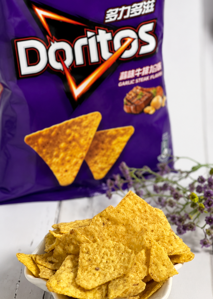 A bag of Doritos Garlic Steak Flavored tortilla chips stands behind a white bowl filled with golden yellow triangular chips on a white wooden surface, with small purple flowers to the right.