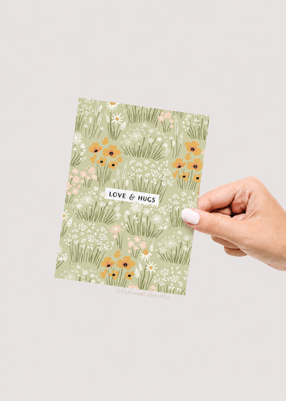 Hand holding a card with floral design and 'Love & Hugs' text on a light gray background