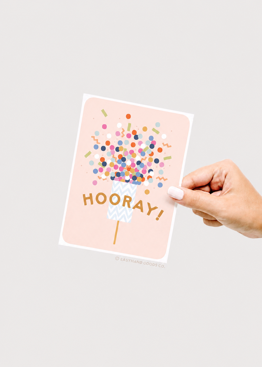 Hand holding a card with 'HOORAY!' and colorful confetti design on a light gray background