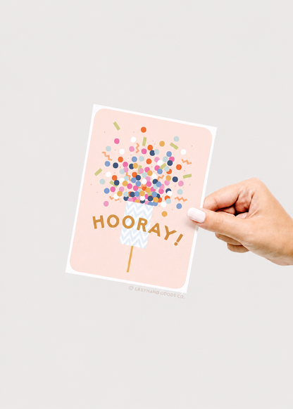 Hand holding a card with 'HOORAY!' and colorful confetti design on a light gray background