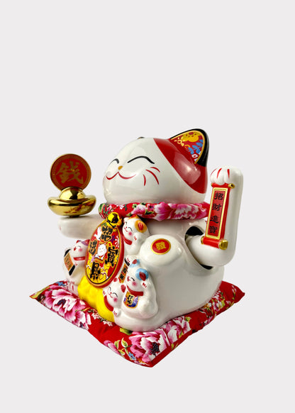 A white ceramic Japanese maneki-neko lucky cat figurine holding a gold ingot and a red scroll, sitting on a red floral cushion. The figurine features traditional Chinese-style gold coins and colorful decorative details, set against a plain white background.