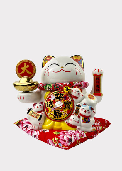 A white ceramic Japanese maneki-neko lucky cat figurine with gold, red, and multicolor floral details, holding a gold ingot and a red vertical plaque, standing on a red floral cushion against a plain white background.