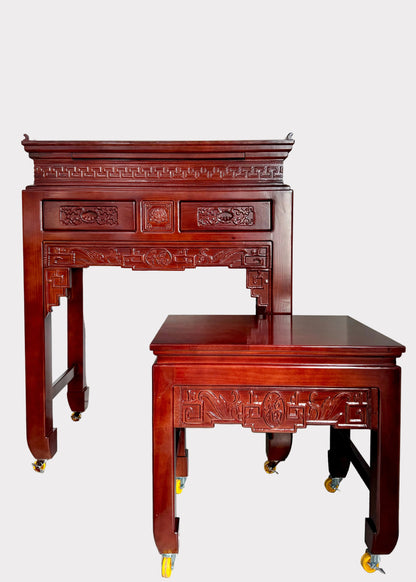 A dark reddish-brown carved wooden writing desk and matching stool set on casters, photographed indoors against a plain light wall and speckled gray floor.