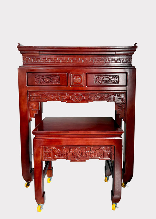 An ornate reddish-brown wooden console table with matching stool, both on casters, photographed in a room corner.