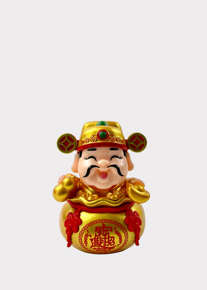 A small decorative figurine of a smiling Chinese good-luck character wearing a gold hat and holding a gold money bag with red details, photographed against a plain light background.
