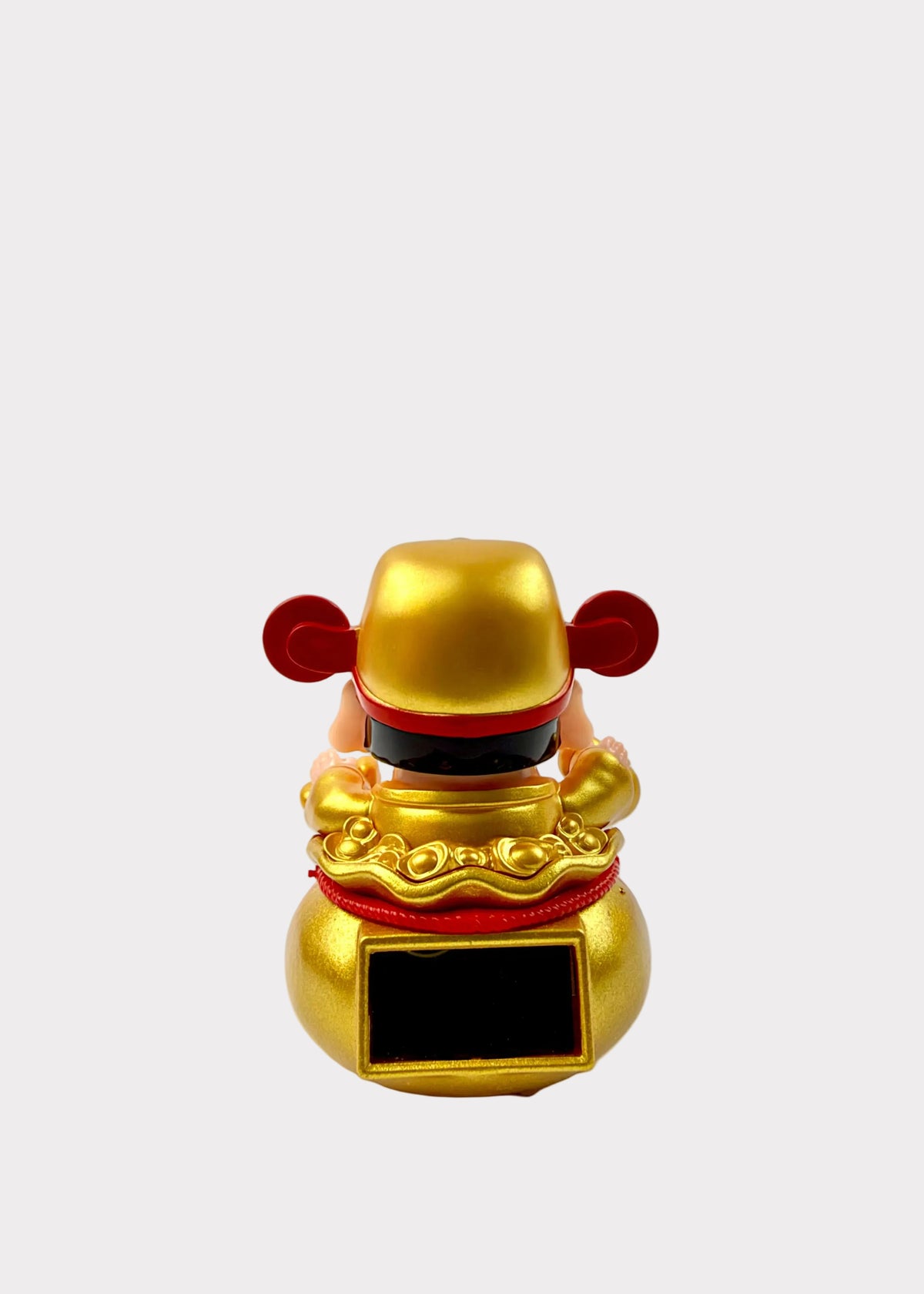 A small golden figurine with red accents and a black rectangular panel on the front, photographed against a plain white wall and white surface.