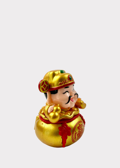A small decorative figurine of a smiling man in traditional Chinese attire sitting on a large gold money bag, photographed on a plain light background.