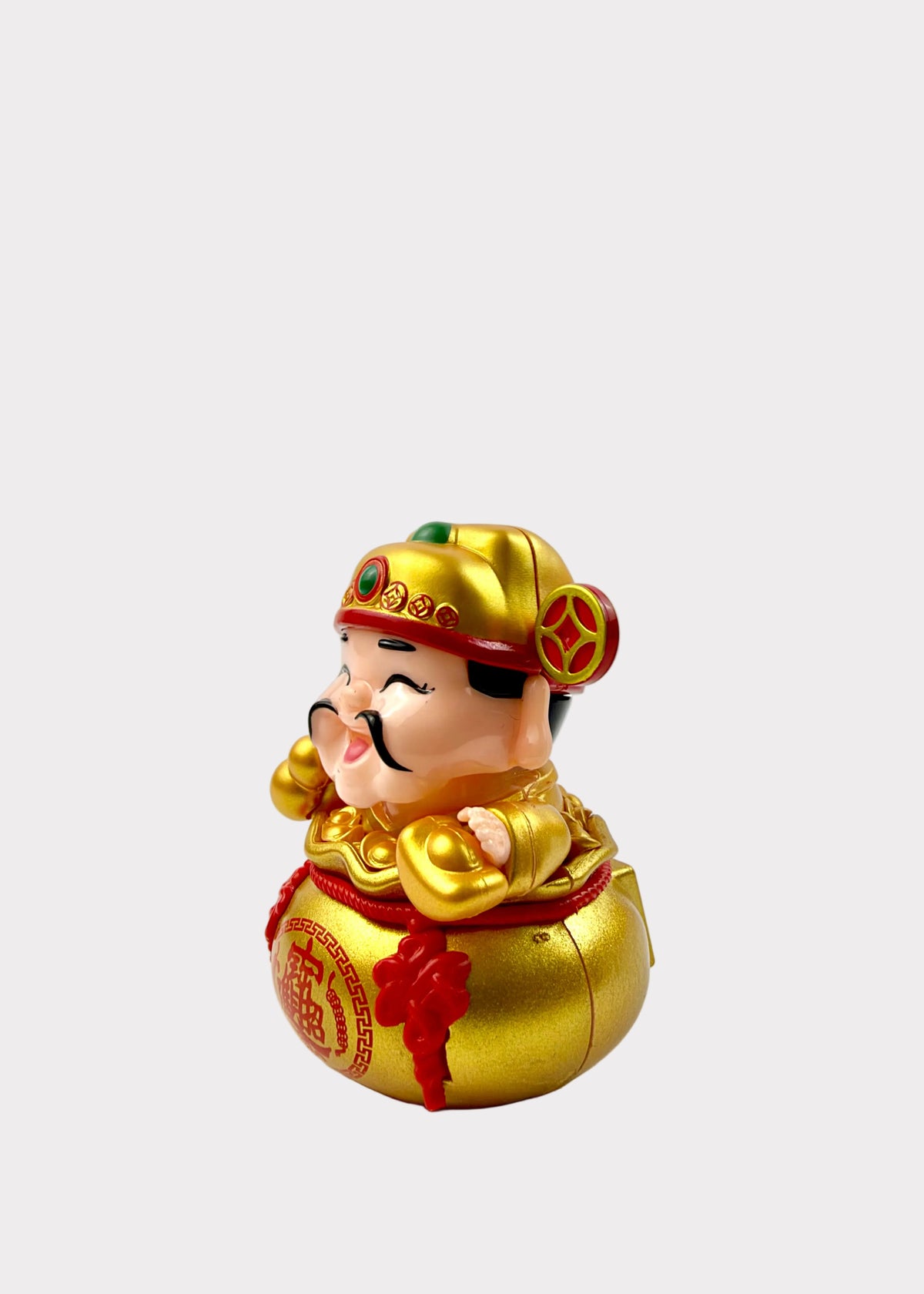 A small decorative figurine of a smiling man wearing a gold hat and robe, seated on a large gold treasure bag with red details, photographed against a plain light background.