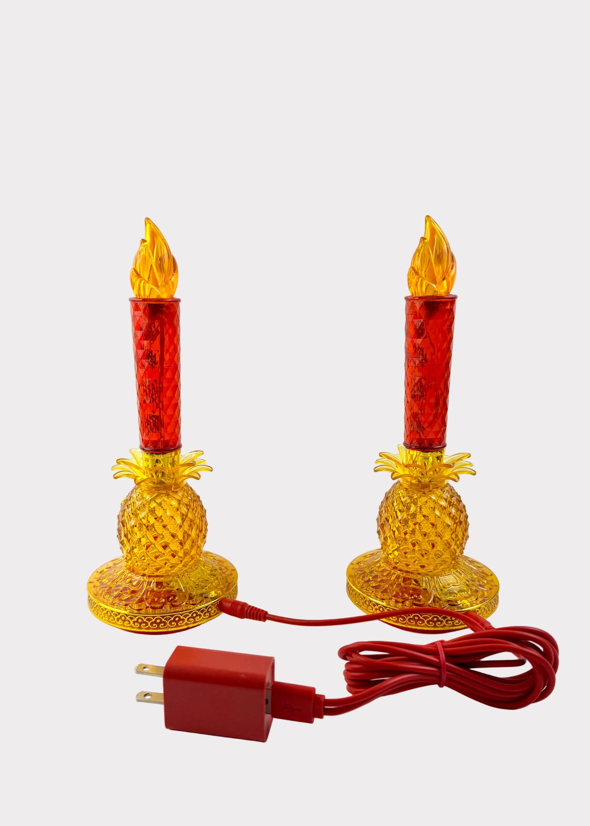 Two decorative candle holders shaped like yellow pineapple bases with red textured candle sleeves and flame tops, shown on a white surface against a white wall, with a red power cord and plug in front.