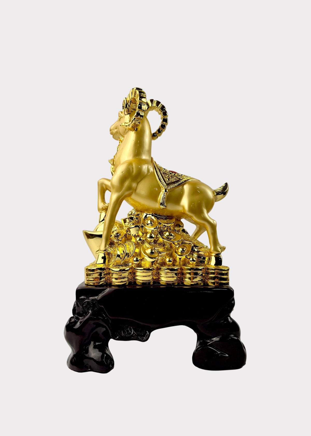 A golden ram figurine standing on a pile of gold coins, mounted on a dark glossy pedestal, photographed against a plain light gray wall and white surface.