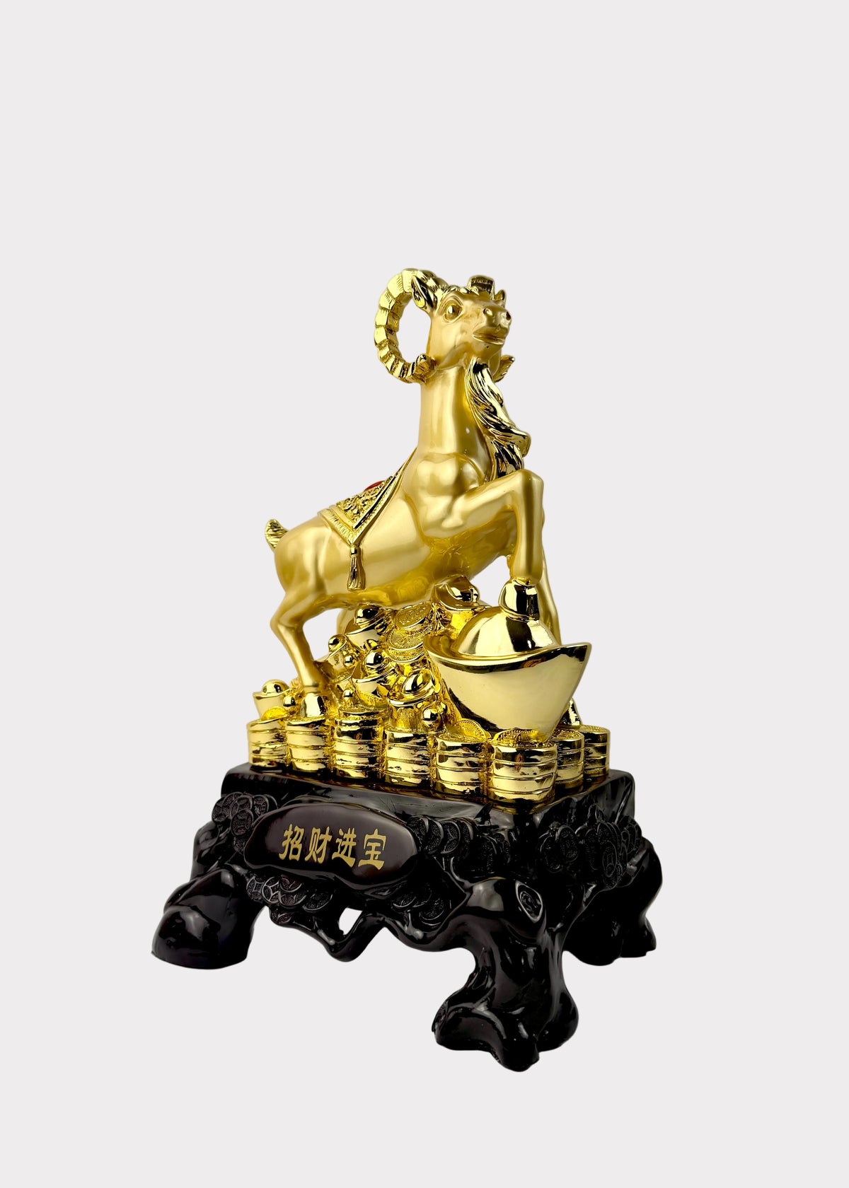 A golden ram figurine standing on a stack of gold ingots and a large gold ingot, mounted on a black decorative base with Chinese characters, photographed against a plain light gray wall and white surface.