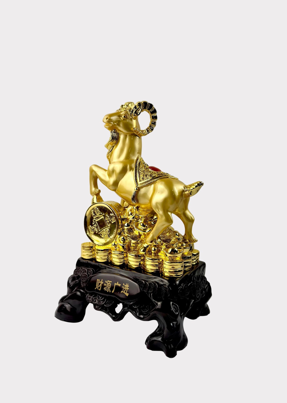 A golden ram figurine standing on a stack of gold coins, mounted on a dark carved base with Chinese characters, photographed against a plain light gray wall and white surface.