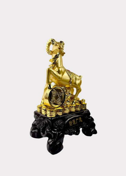 A golden ram figurine standing on a stack of gold coins, mounted on a black carved base with Chinese characters, photographed against a plain light gray wall and white surface.