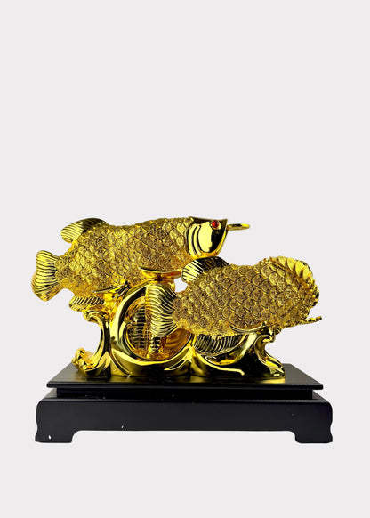 A golden fish sculpture with detailed scales and a small red eye accent, mounted on a black rectangular base against a plain light gray wall.