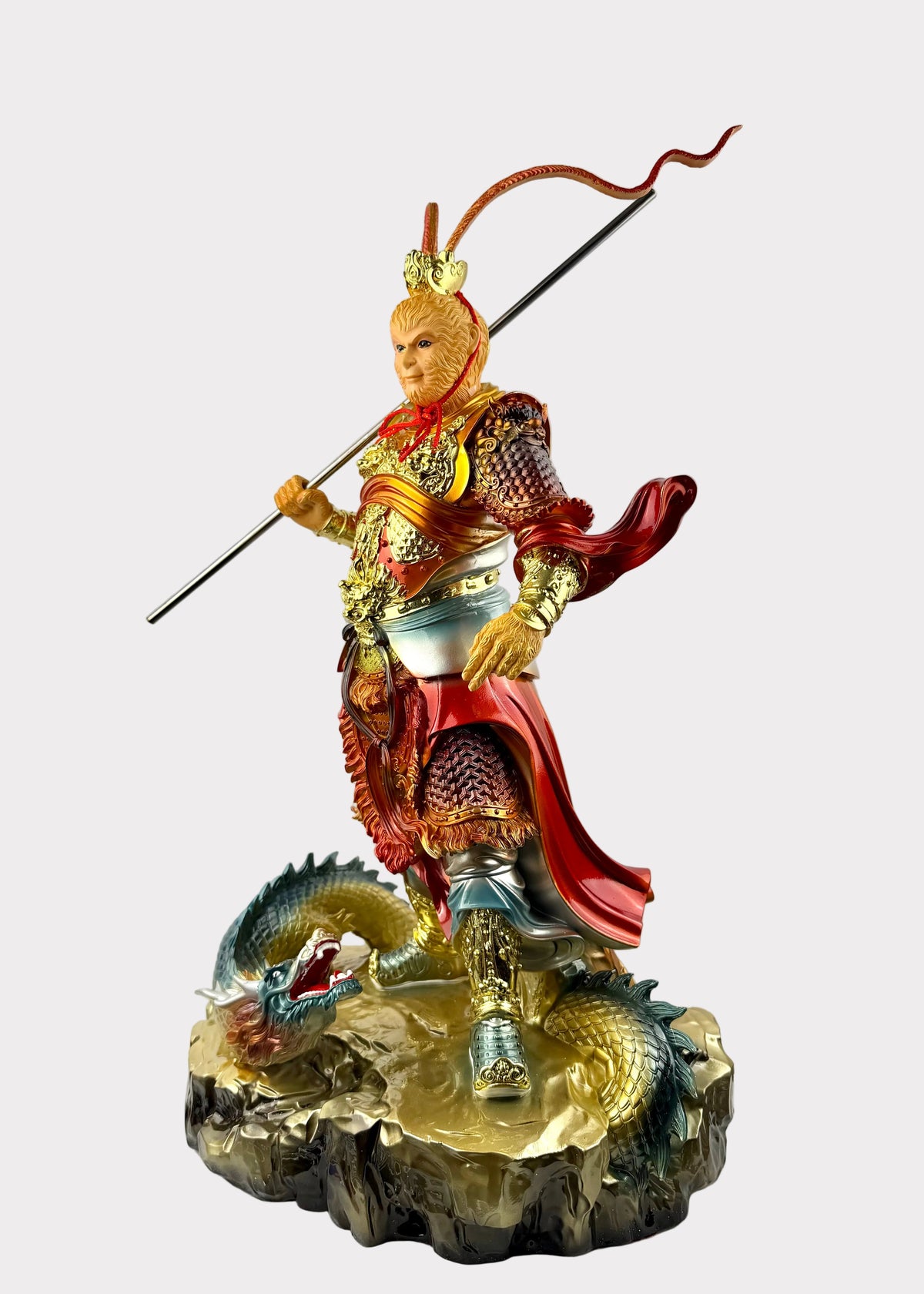 A detailed figurine of a monkey warrior in ornate red and gold armor, holding a long spear, standing on a rocky base with a green dragon at its feet. The figurine is photographed indoors against a plain light wall, resting on a white sheet over a wire shelf.