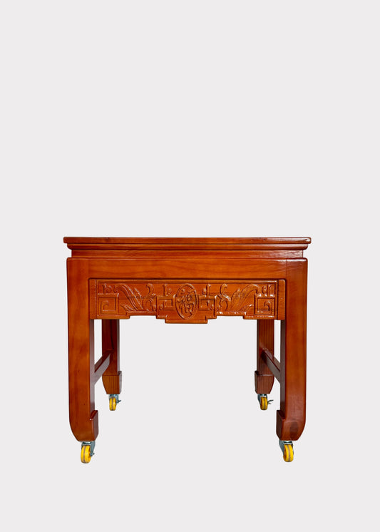 A reddish-brown wooden side table with ornate carved details on the front panel and legs, supported by four casters, set against a plain off-white wall and speckled gray floor.