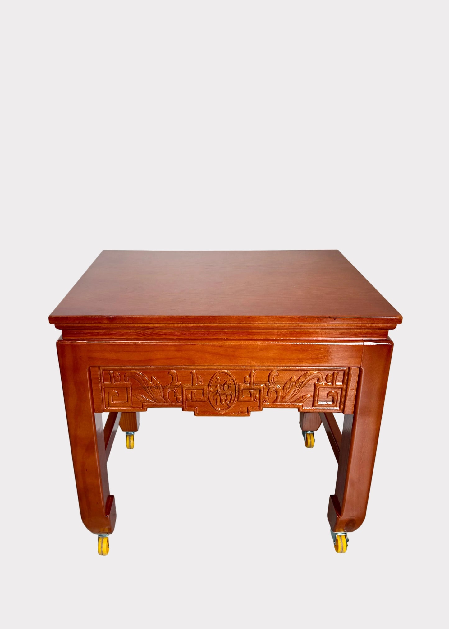 A reddish-brown wooden rolling side table with ornate carved details on the front panel and yellow caster wheels, photographed against a plain light gray wall and speckled gray floor.