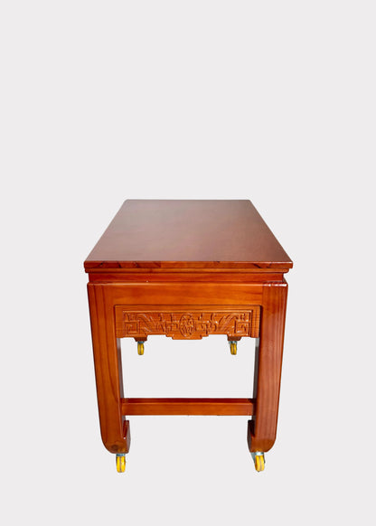 A reddish-brown wooden rolling side table or cart with a carved front panel and four caster wheels, photographed against a plain light wall and speckled gray floor.