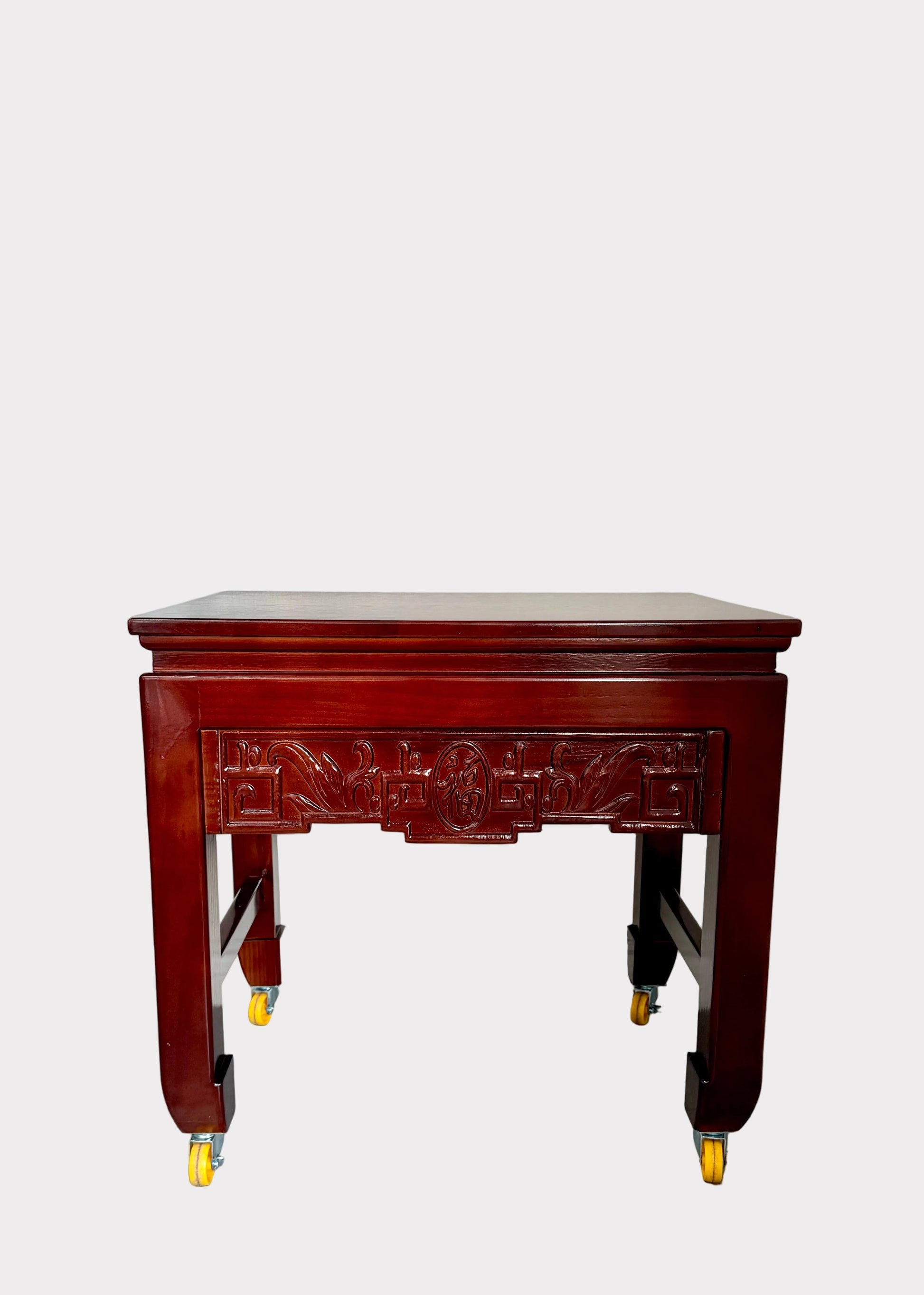 A dark reddish-brown wooden rolling side table with ornate carved details on the front panel and legs, fitted with yellow caster wheels, set against a plain white wall and gray speckled floor.