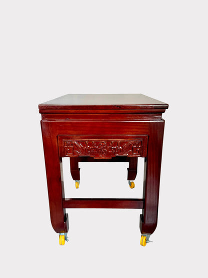 A dark reddish-brown wooden rolling side table with a carved drawer front, four legs, and yellow caster wheels, photographed in a room corner against a white wall and speckled gray floor.