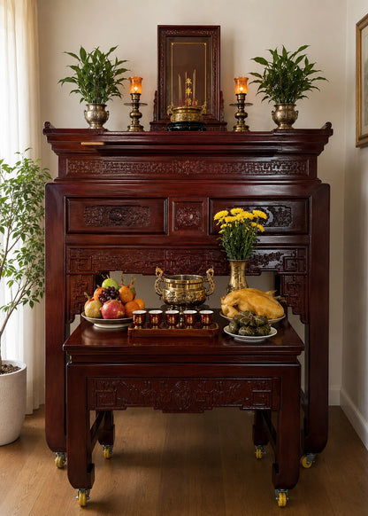 An ornate dark reddish-brown carved wooden altar cabinet with casters, styled with potted plants, candles, fruit, flowers, and a whole roasted bird in an indoor room.