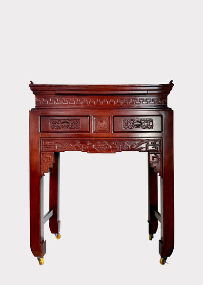 A traditional carved wooden console table with a reddish-brown finish and gold caster wheels, set against a plain white wall.