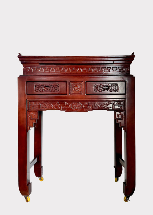 A traditional carved wooden console table with a reddish-brown finish and gold caster wheels, set against a plain white wall.