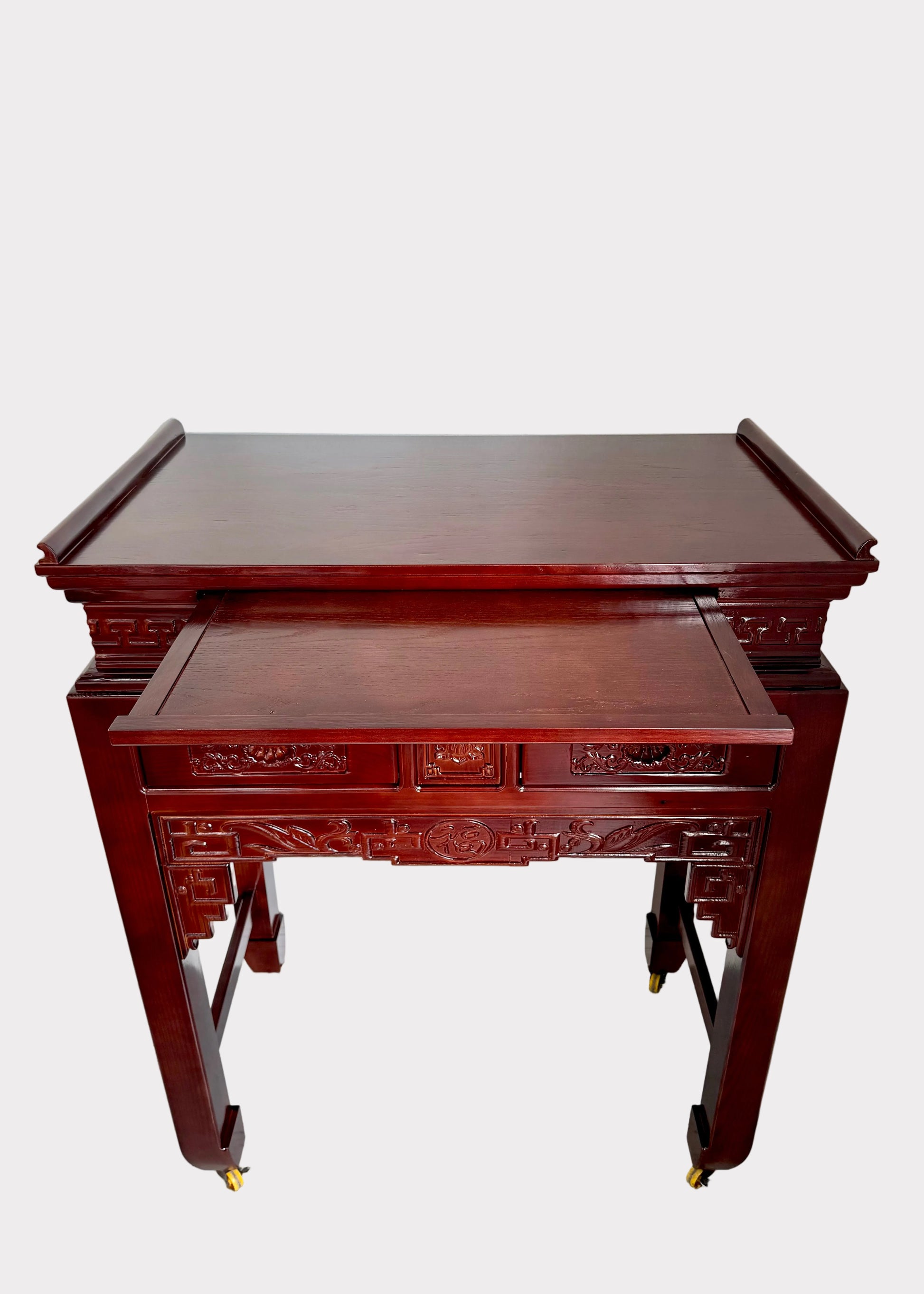 A dark reddish-brown wooden console table with ornate carvings and casters, photographed in a room corner against a white wall and speckled gray floor.