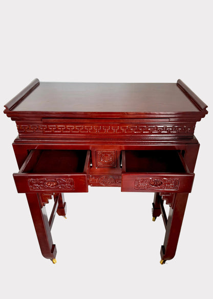 A dark reddish-brown wooden writing desk with carved details and two open drawers, photographed in a room corner against a white wall and speckled gray floor.