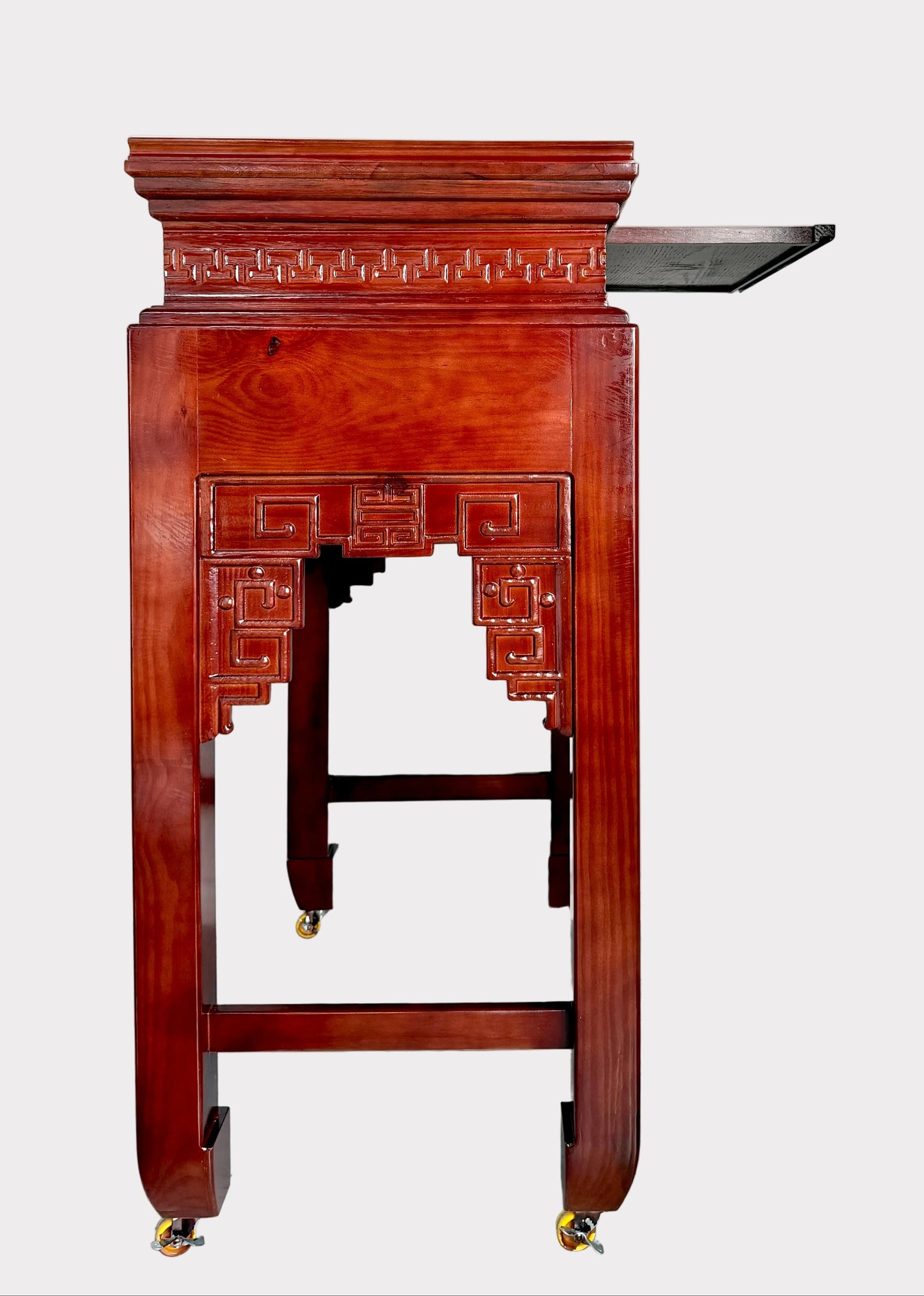 A reddish-brown wooden display stand or credenza with ornate carved details and small brass caster wheels, photographed in a room with white walls and gray floor.
