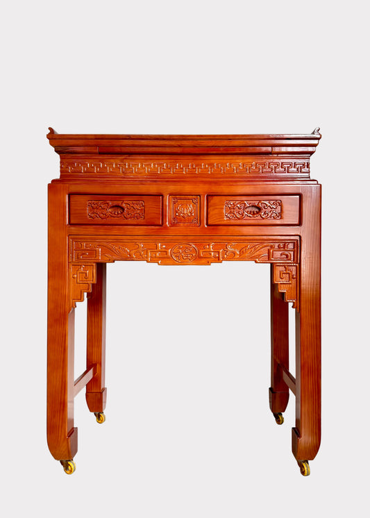 A traditional Chinese red wood console table with ornate carvings and brass casters, photographed against a plain white wall and gray speckled floor.