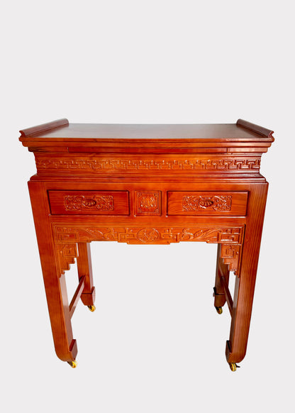 A traditional Chinese wooden console table with ornate carvings, two drawers, and four legs on small brass casters, shown against a plain white wall and gray speckled floor.