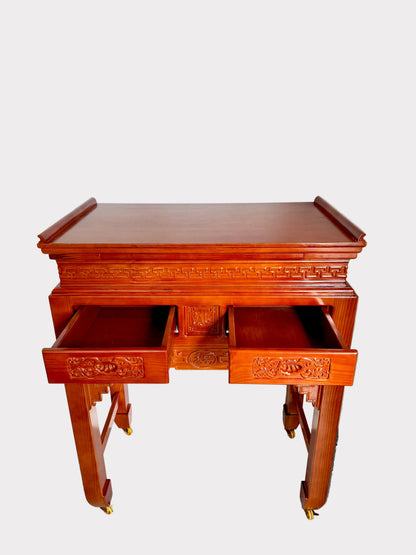 A reddish-brown wooden writing desk with carved details and two open drawers, photographed against a plain white wall and gray speckled floor.