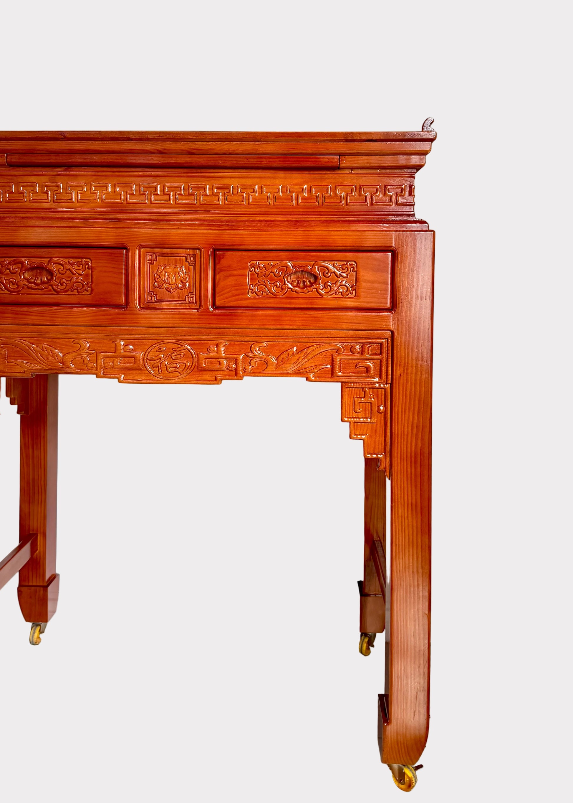 A traditional Chinese-style wooden console table with ornate carvings and brass caster wheels, positioned against a plain white wall on a speckled gray floor.