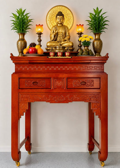 A carved reddish-brown wooden altar table with gold-tone caster wheels, styled with a golden Buddha statue, lotus candles, potted plants, and fruit against a plain light gray wall.