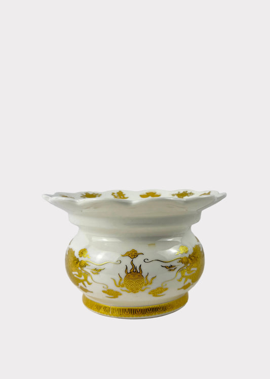A small white ceramic cup or candle holder with a flared rim, decorated with gold floral and scroll motifs, set against a plain light gray background.