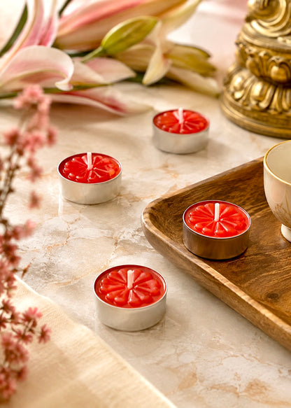 Four small red tea light candles in silver metal holders arranged on a marble surface with decorative flowers, a wooden tray, and a gold-toned object in the background.