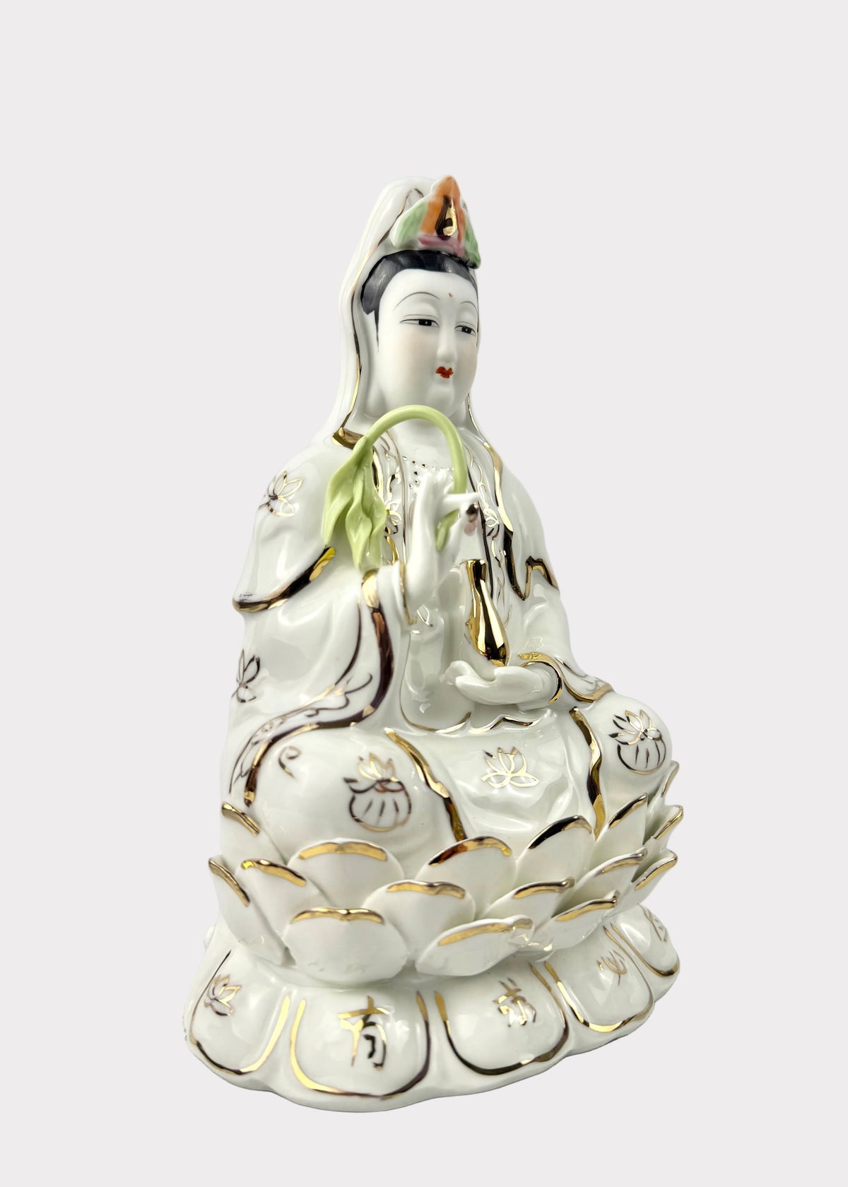 A white porcelain figurine of Guanyin seated on a lotus, decorated with gold accents and green leaf details, set against a plain light gray background.