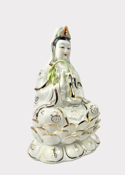 A white porcelain figurine of Guanyin seated on a lotus, decorated with gold accents and green leaf details, set against a plain light gray background.