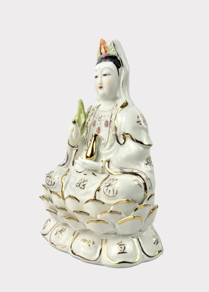 A white porcelain Guanyin statue seated on a lotus base, decorated with gold trim and small multicolor accents, photographed against a plain light gray background.