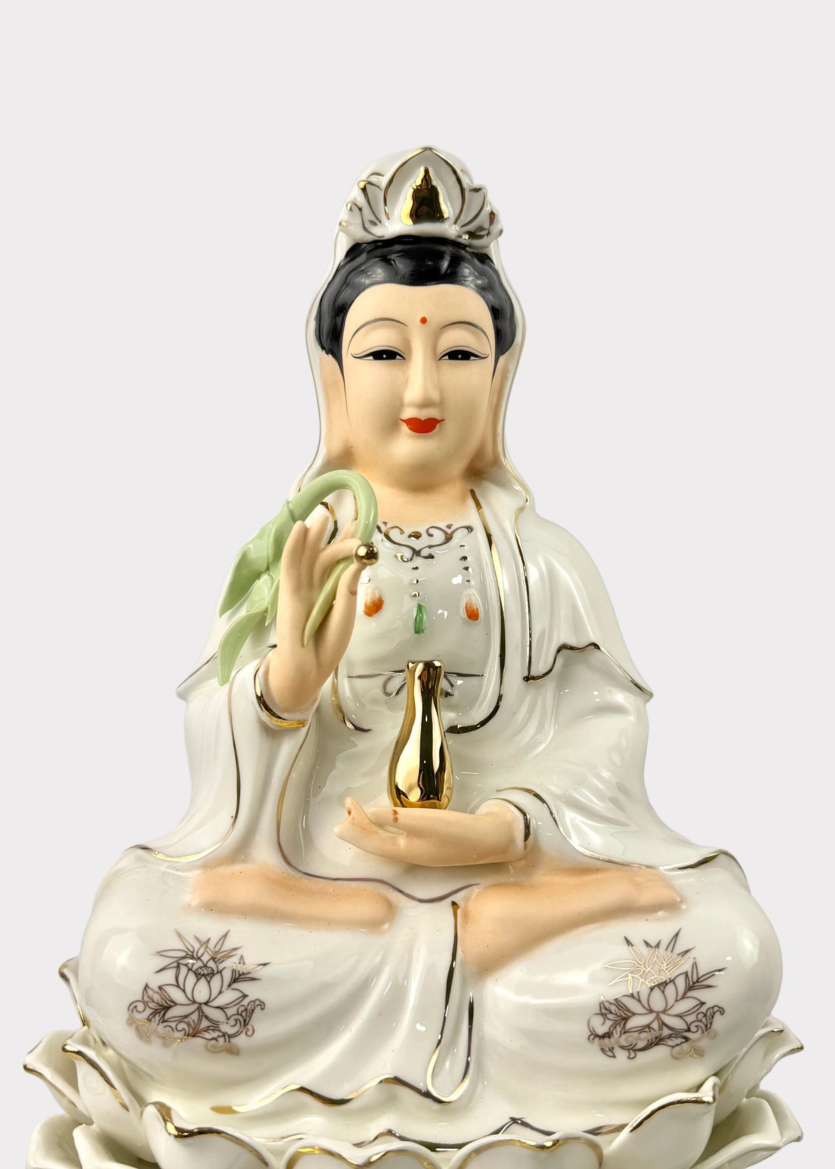 A ceramic figurine of a seated female deity with black hair, wearing a white robe with gold trim and floral motifs, holding a green lotus and a gold vessel.