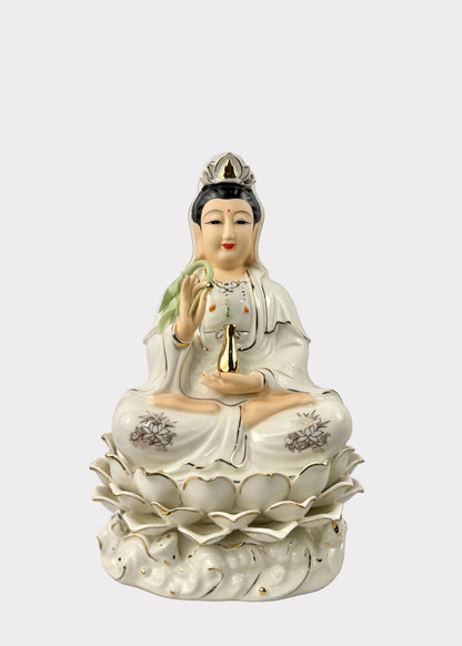 A white porcelain figurine of a seated female deity with black hair, wearing a crown and robe, holding a small gold vessel, displayed on a lotus base against a plain light gray background.